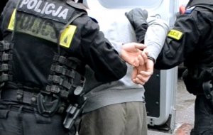 Na zdjęciu dwóch policjantów i mężczyzna w kajdankach zapiętych na ręce z tyłu. Stoją przy radiowozie.