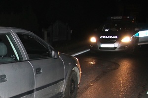 Na zdjęciu noc na ulicy stoi radiowóz z włączonymi światłami oraz samochód osobowy