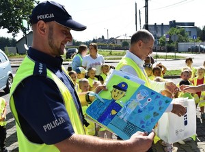 policjant - sołtys prezentuje  obrazek namalowany przez przedszkolaków, w tle wójt i dzieci