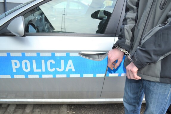 Na zdjęciu mężczyzna w kajdankach na tle radiowozu