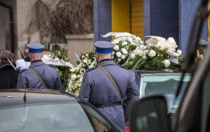 Na zdjęciu samochód typu karawan z wieńcami na dachu. Za nim policjanci z bronią.