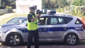 Na zdjęciu policjant stojący na ulicy. W dłoni trzyma miernik prędkości. W obok stoi radiowóz.