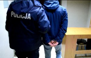 Na zdjęciu policjant oraz stojący obok mężczyzna w kajdankach zapiętych na ręce z tyłu.