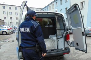 Na zdjęciu policjant obok radiowozu. Radiowóz ma otwarte tylne drzwi z kratą.