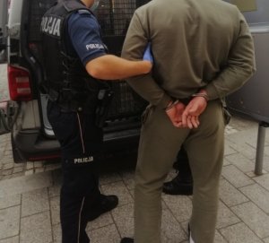 Na zdjęciu policjant, a  obok stoi zatrzymany mężczyzna z kajdankami zapiętymi na ręce z tyłu. Są przy radiowozie.