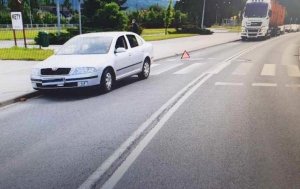 Na zdjęciu ulica na której stoi samochód. Pojazd stoi w rejonie przejścia dla pieszych. W tle ulicą jedzie ciężarówka.