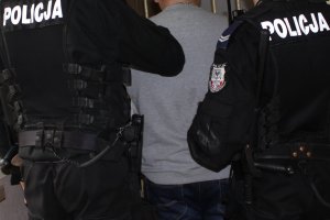 Na zdjęciu mężczyzna przed kratami obok dwaj umundurowani policjanci