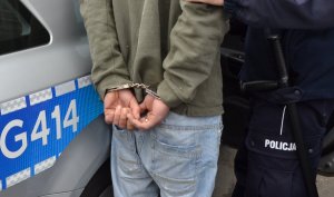 Na zdjęciu zatrzymany z kajdankami zapiętymi na ręce z tyłu obok stoi policjant