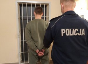 Na zdjęciu pomieszczenie. Policjant wraz z zatrzymanym stoją przed drzwiami z kratą.