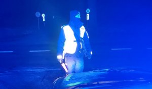 Na zdjęciu policjant pełniący służbę w nocy na drodze. Obok stoi radiowóz z włączonymi światłami.