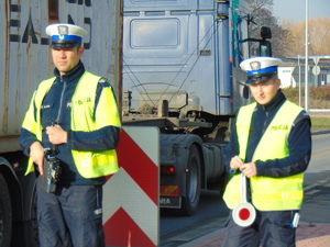 Na zdjęciu dwóch policjantów stoi na drodze. W tle  jadące ciężarówki.