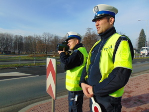Dwóch policjantów stoi przy drodze. Jeden w ręku trzyma miernik i dokonuje pomiaru prędkości.