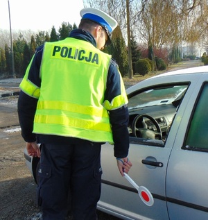 Na zdjęciu policjant drogówki stojący przy samochodzie. Policjant w ręku ma tarczę do zatrzymywania pojazdów.