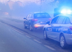 Na zdjęciu radiowóz policyjny z włączonymi sygnałami na ulicy. Przed radiowozem stoi samochód osobowy.