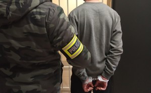 Na zdjęciu policjant w ubraniu cywilnym z opaską Policja na ramieniu. Obok zatrzymany w kajdankach zapiętych na ręce z tyłu.