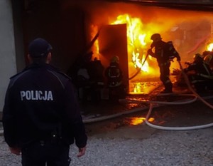 Na zdjęciu pożar. Strażak gasi ogień w budynku. Policjant pilnuje bezpieczeństwa na zewnątrz.