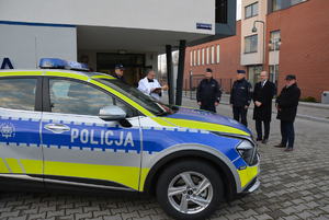 Na zdjęciu policjanci i osoby cywilne stoją przed budynkiem komisariatu Policji. Obok stoi nowy oznakowany radiowóz.
